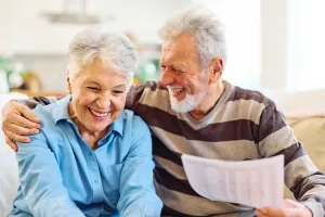 Smiling senior couple sitting together at home, with the man holding paperwork and his arm around the woman.